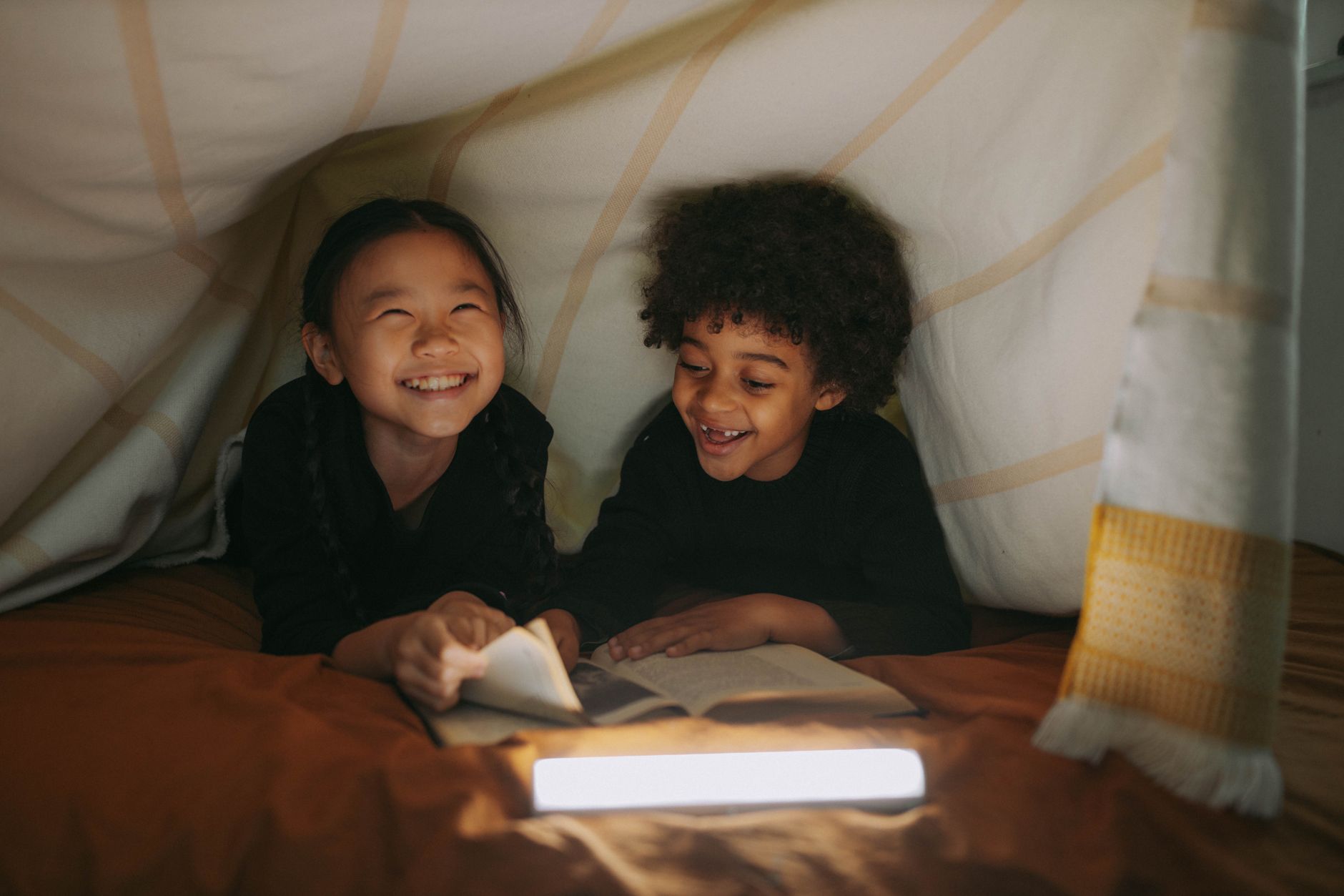Deux enfants souriants lisent un livre dans une cabane en couvertures